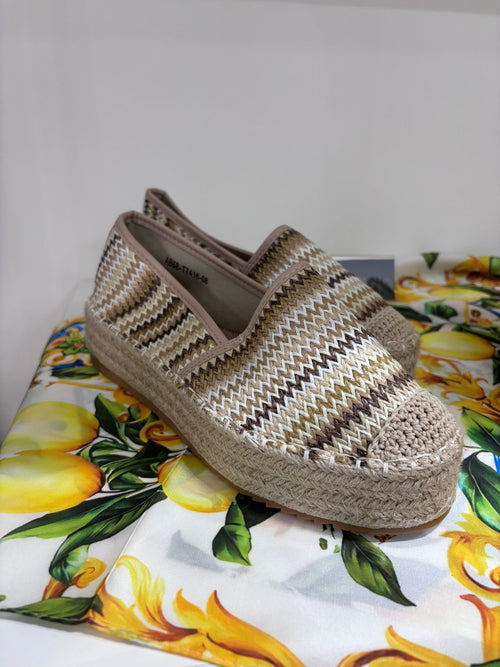 Slip-on espadrillas with beige, brown, and white woven pattern and braided jute sole on lemon-patterned fabric background.