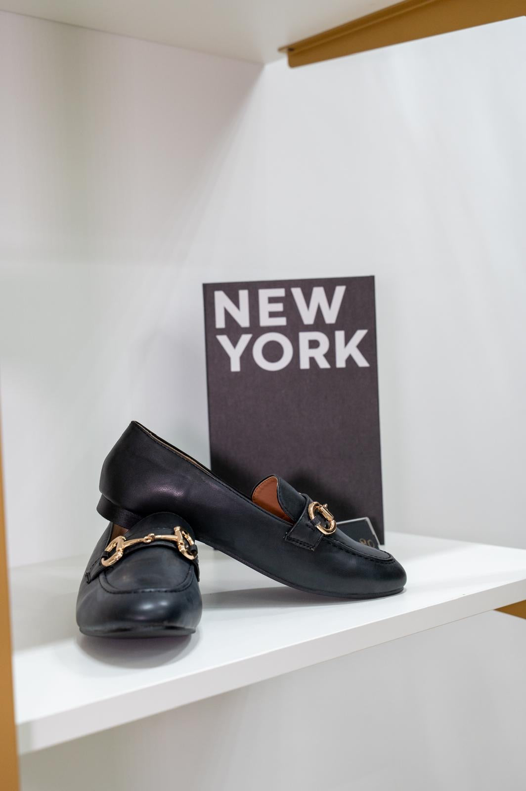 Black leather moccasin loafers with gold-tone horsebit detail and rounded toe on white shelf.