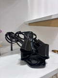 Black glossy high-heeled sandal with square toe and large decorative bow on front strap.