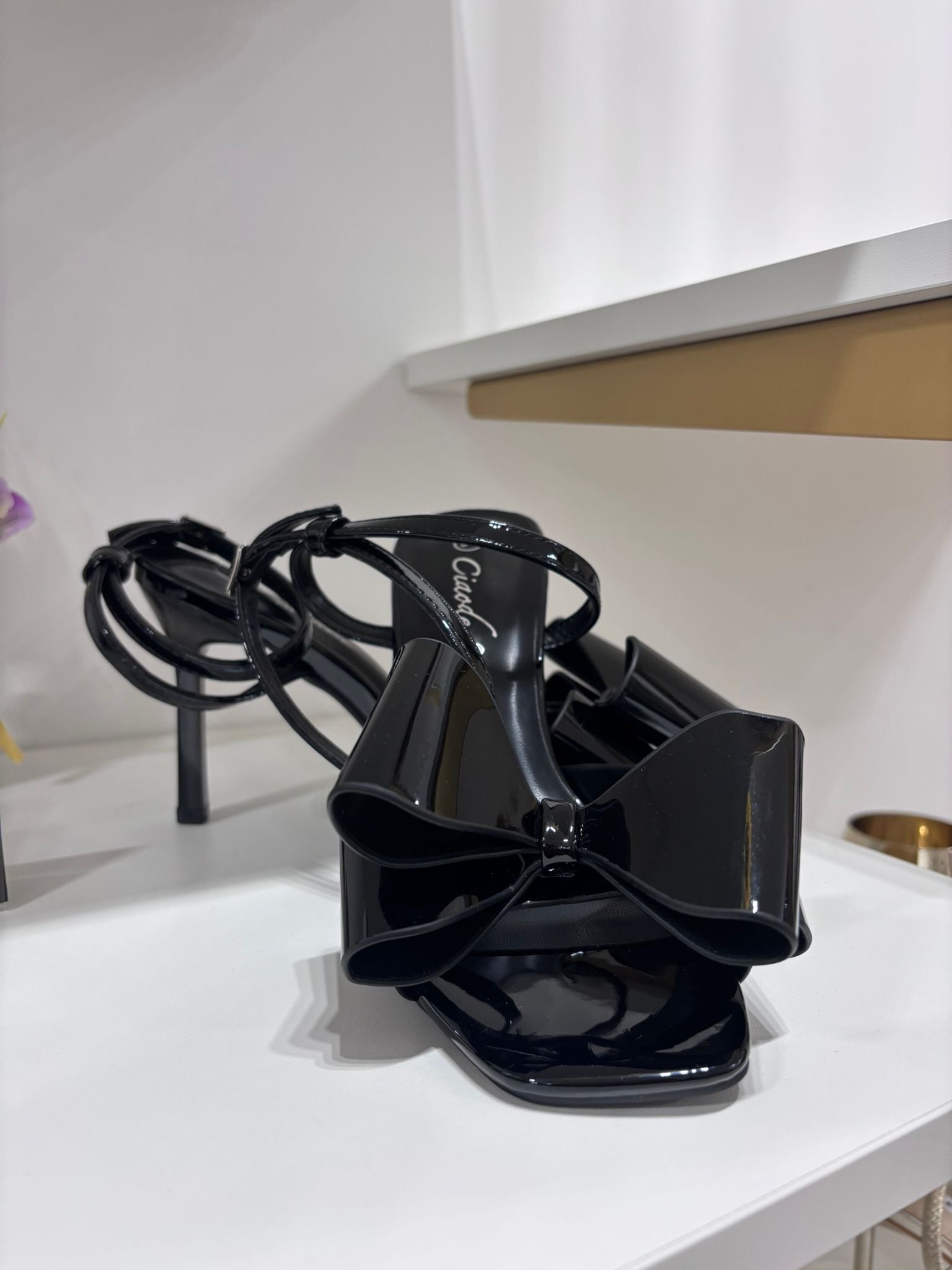Black glossy high-heeled sandal with square toe and large decorative bow on front strap.