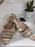Beige woven straw slingback flats with pointed toes and adjustable ankle straps shown with potted plant and black box.