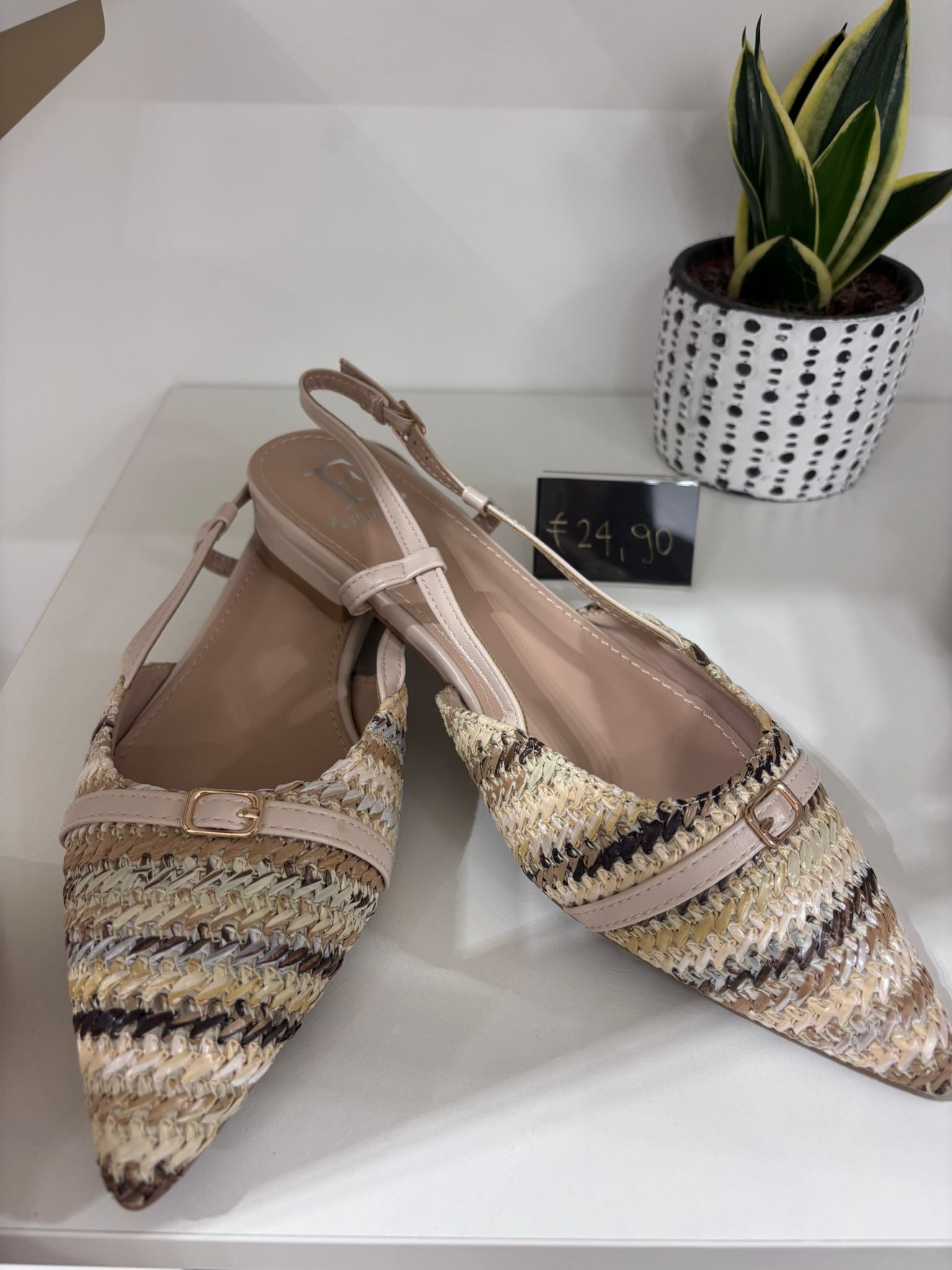 Beige woven straw slingback flats with pointed toes and adjustable ankle straps shown with potted plant and black box.