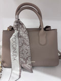 Taupe leather handbag with rounded handles and a decorative brown patterned silk scarf tied around one handle.