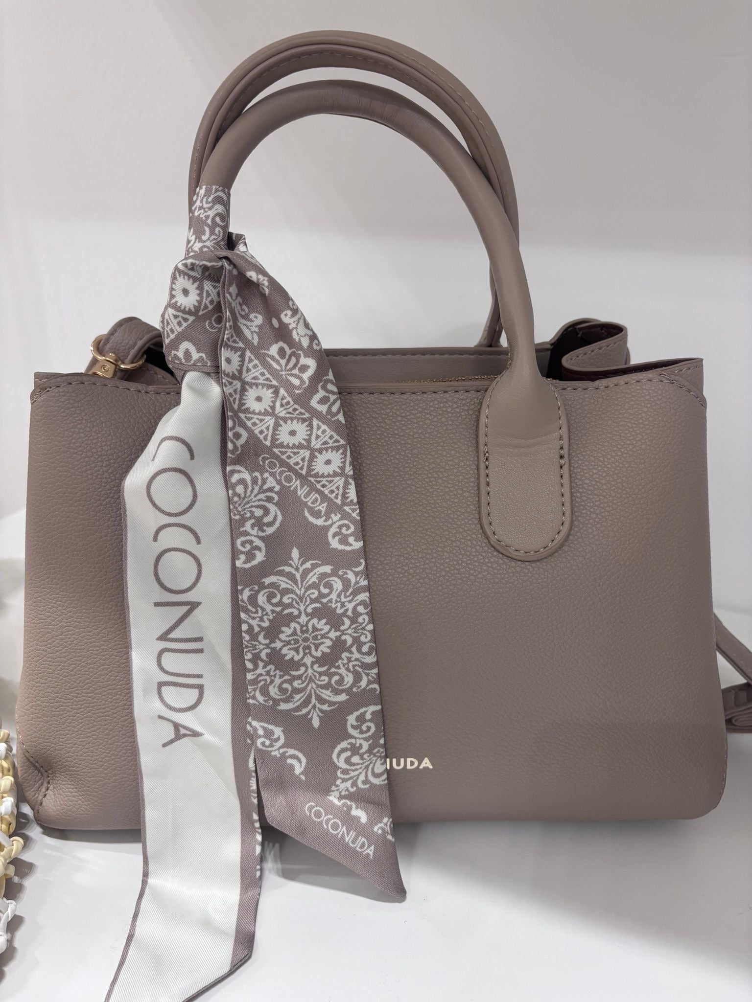 Taupe leather handbag with rounded handles and a decorative brown patterned silk scarf tied around one handle.