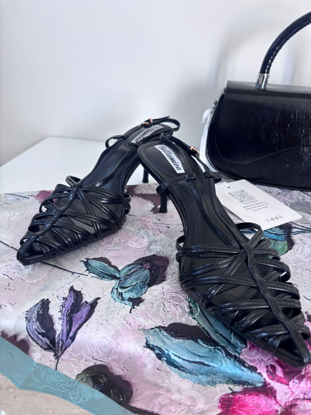 Black slingback sandals with woven leather straps and low block heels on floral surface with black handbag in background.