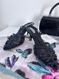 Black slingback sandals with woven leather straps and low block heels on floral surface with black handbag in background.