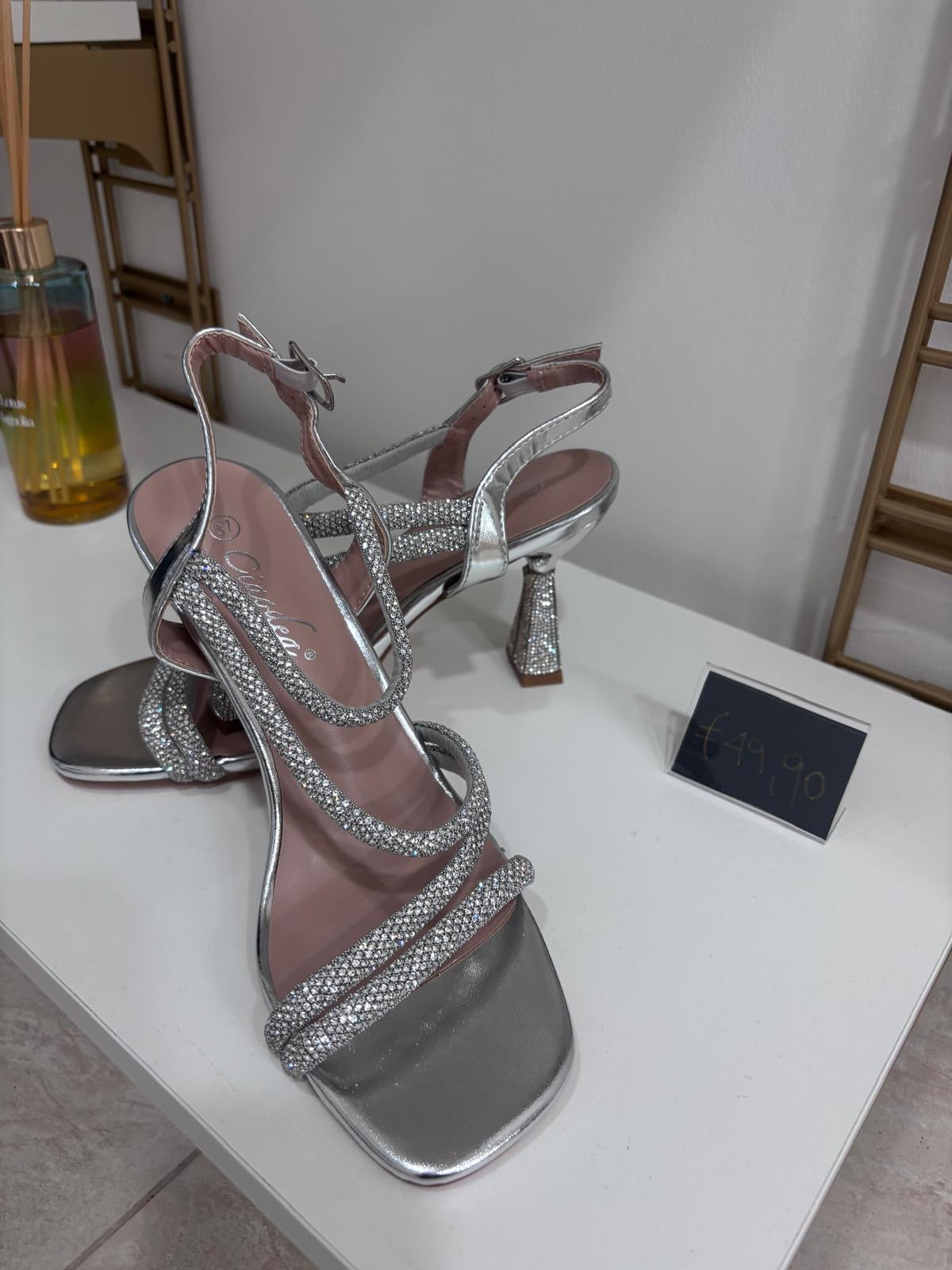 Silver strappy high-heeled sandals with double crystal embellishments and ankle straps on a white surface.
