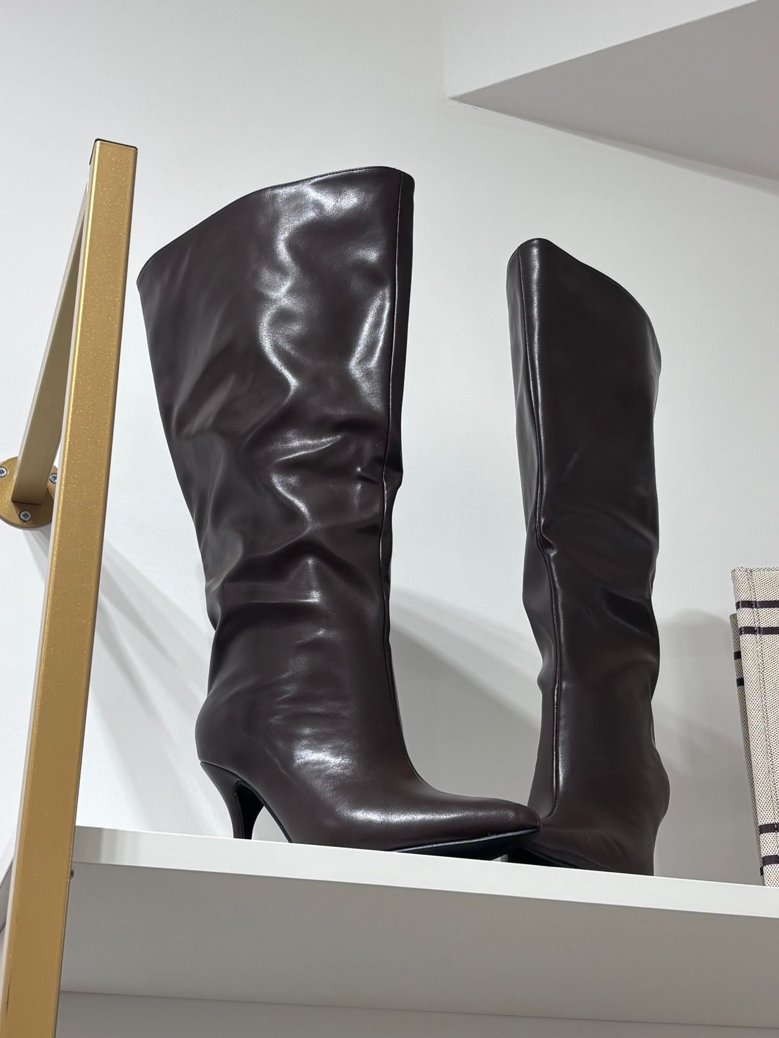 Tall brown leather boots with a soft, slightly slouched shaft and slim heels on a white shelf.