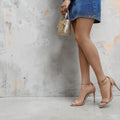 Nude high-heeled sandals with thin ankle straps paired with a denim mini skirt and clear handle handbag against gray.