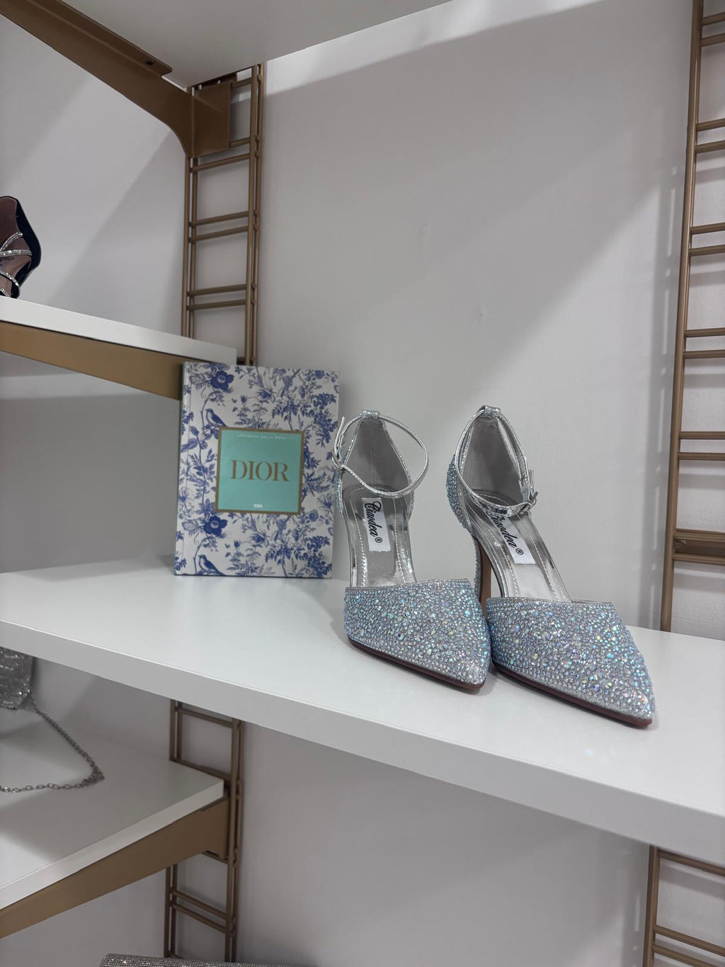 Silver glittery pointed toe high heel décolleté shoes with ankle straps on white shelf.