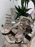 Gold high-heeled sandals with multiple thin straps and buckle details displayed beside a metallic gold pouch on a mirrored.