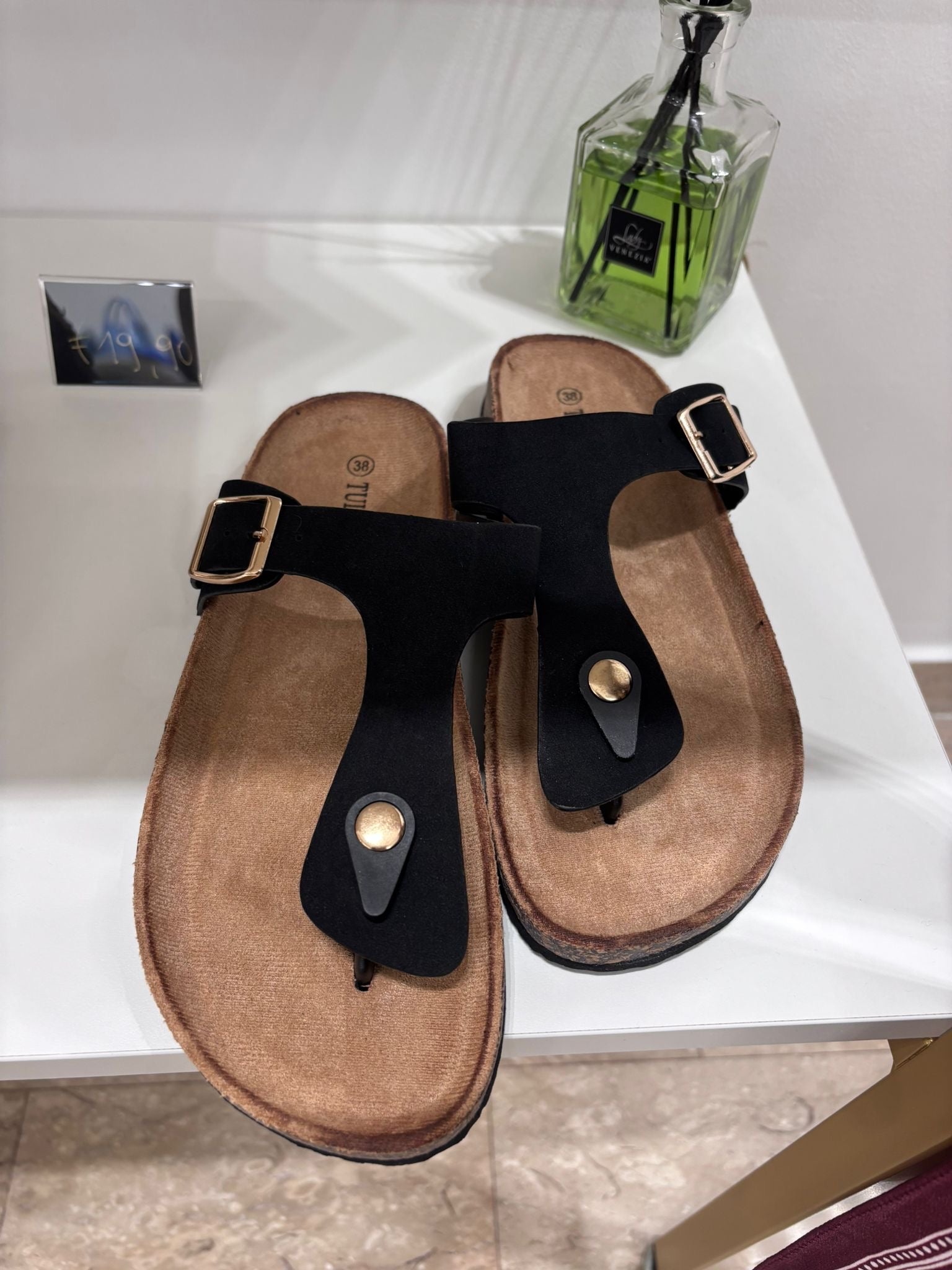Black flat thong sandals with wide straps and gold buckles on tan footbed.